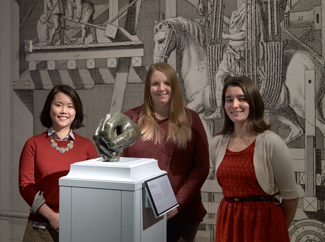 Three young women standing in a museum gallery behind an over-life-size bronze male hand on a pedestal