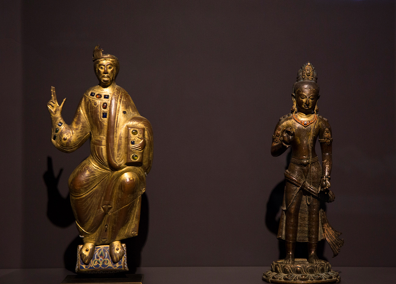 Two small metal devotional sculptures side by side in case, both with their right arms raised in blessing